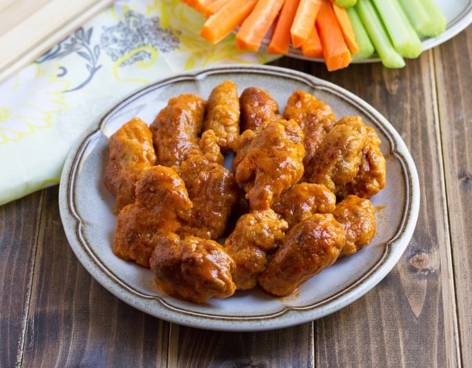 vegan wings on a white plate with brown strips around the edge; cream colored cloth with blue flowers in background and plate with carrot and celery sticks in background
