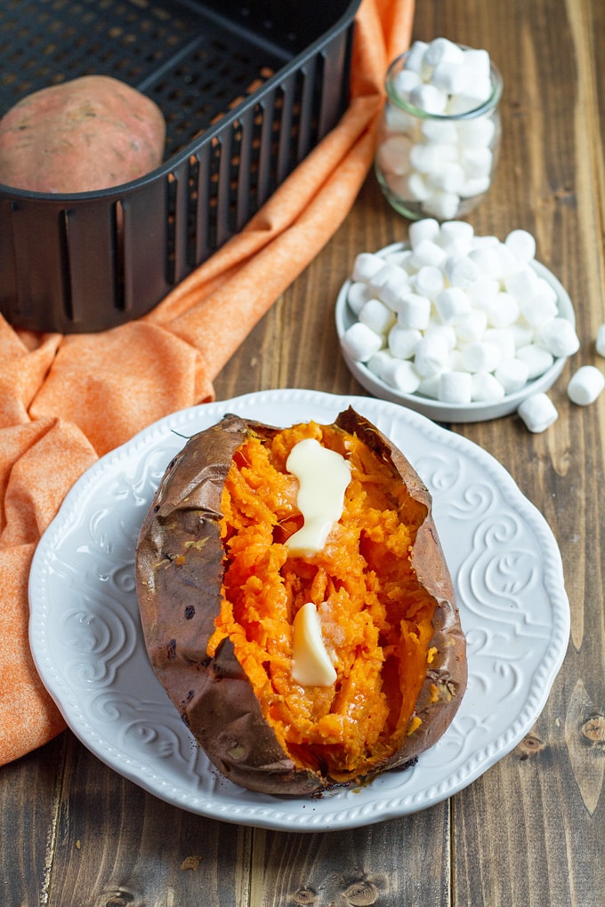 baked sweet potato cut open with butter on a white plate; light orange vloth in background with air fryer basket and small bowl of mini marshmallows