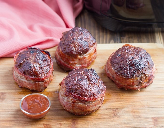 4 mini bacon wrapped meatloaves on wooden cutting board with pinch bowl of ketchup in front; pink cloth in background