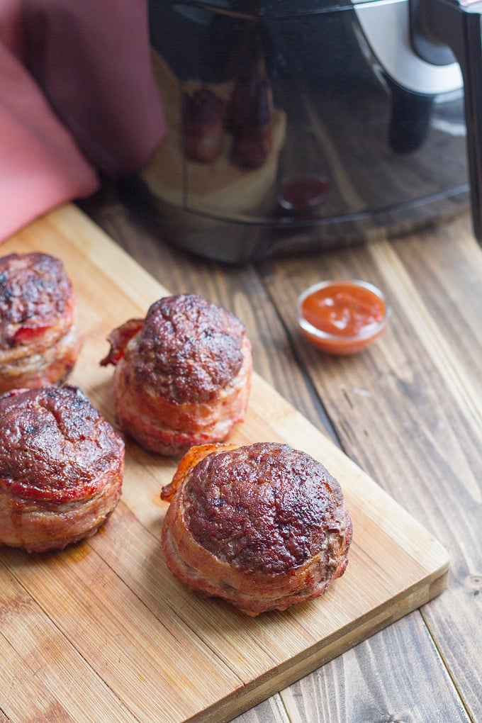 4 mini bacon wrapped meatloaves on wooden cutting board with pinch bowl of ketchup in front; air fryer in background