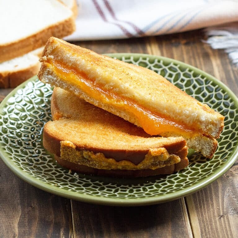 green plate with grilled cheese with orange melty cheese; stack of white bread in background; plaid cloth and air fryer