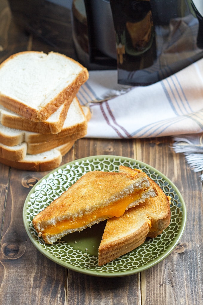 green plate with grilled cheese with orange melty cheese; stack of white bread in background; plaid cloth and air fryer
