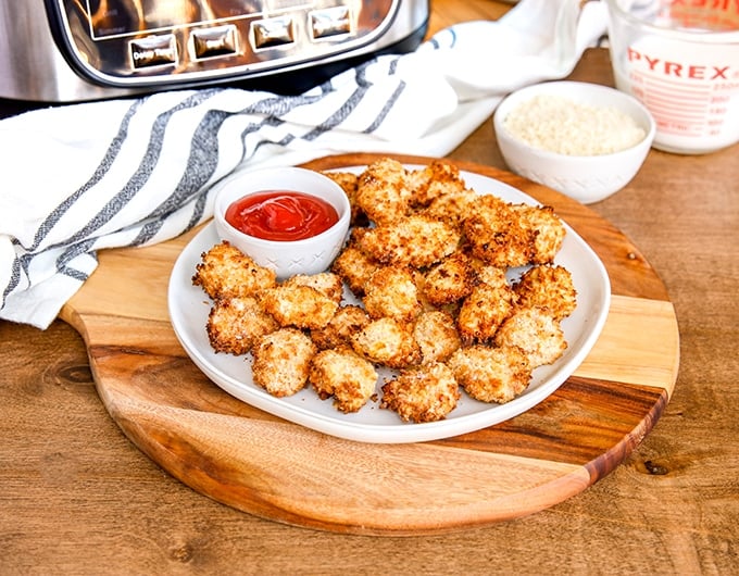 breaded chicken bites on white plate on top wooden server; small white bowl ketchup on plate; white condiment bowl and glass measuring cup to back right, and white cloth with black stripe to back left; bottom of air fryer in top corner
