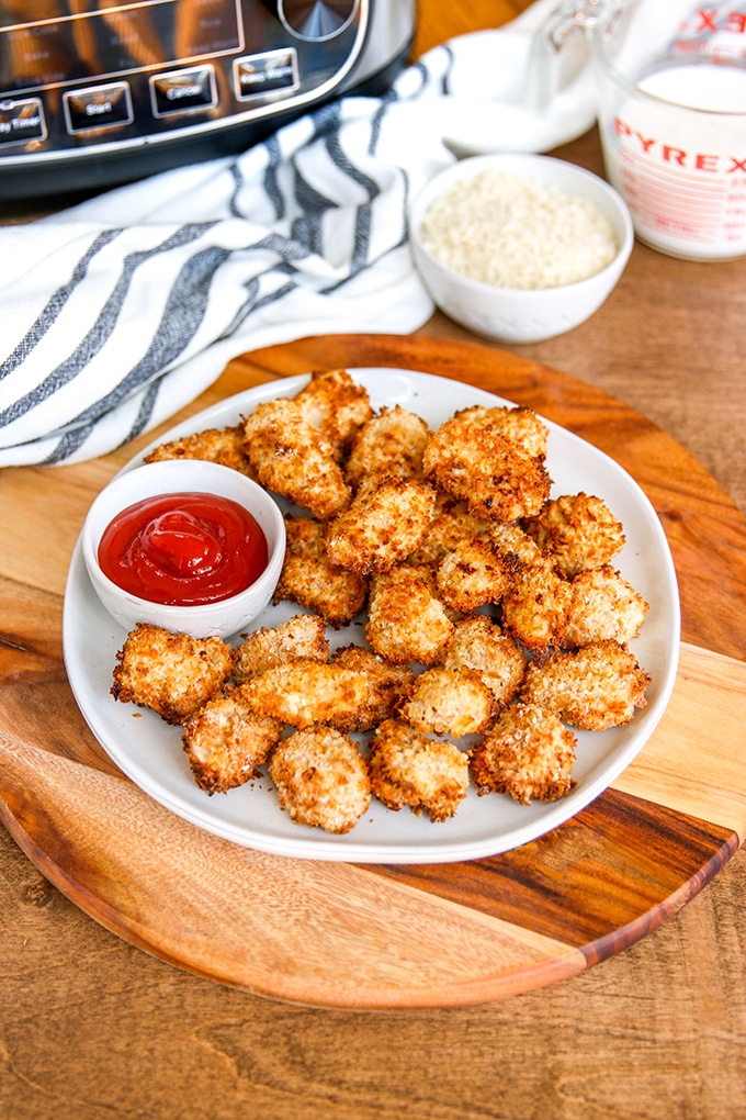 breaded chicken bites on white plate on top wooden server; small white bowl ketchup on plate; white condiment bowl and glass measuring cup to back right, and white cloth with black stripe to back left; bottom of air fryer in top corner