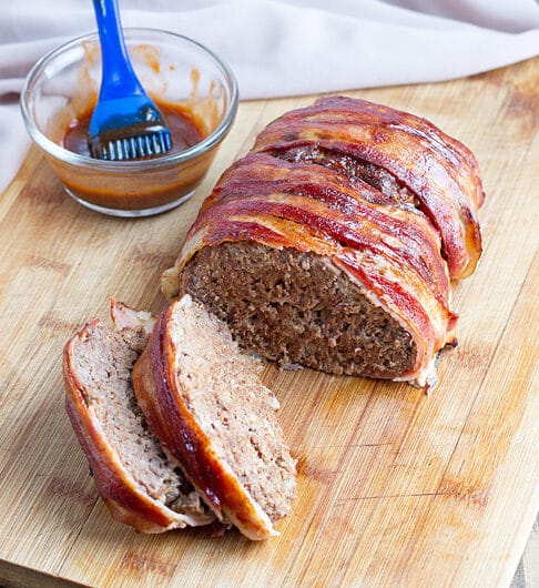 bacon wrapped meatloaf with 2 slices on wooden cutting board; bbq sauce in condiment jar with basting brush in background