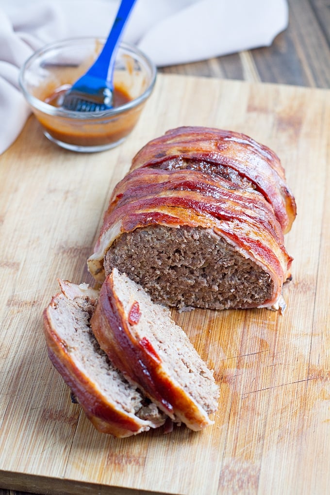 bacon wrapped meatloaf with 2 slices on wooden cutting board; bbq sauce in condiment jar with basting brush in background