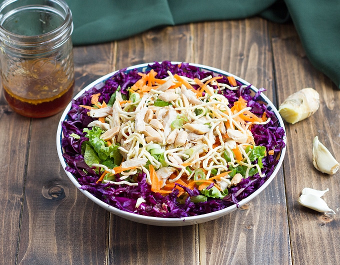 bowl with purple cabbage, lettuce, chicken, peanuts, carrots, crunchy rice noodles; garlic, ginger, and dressing in a jar in background; green cloth in background