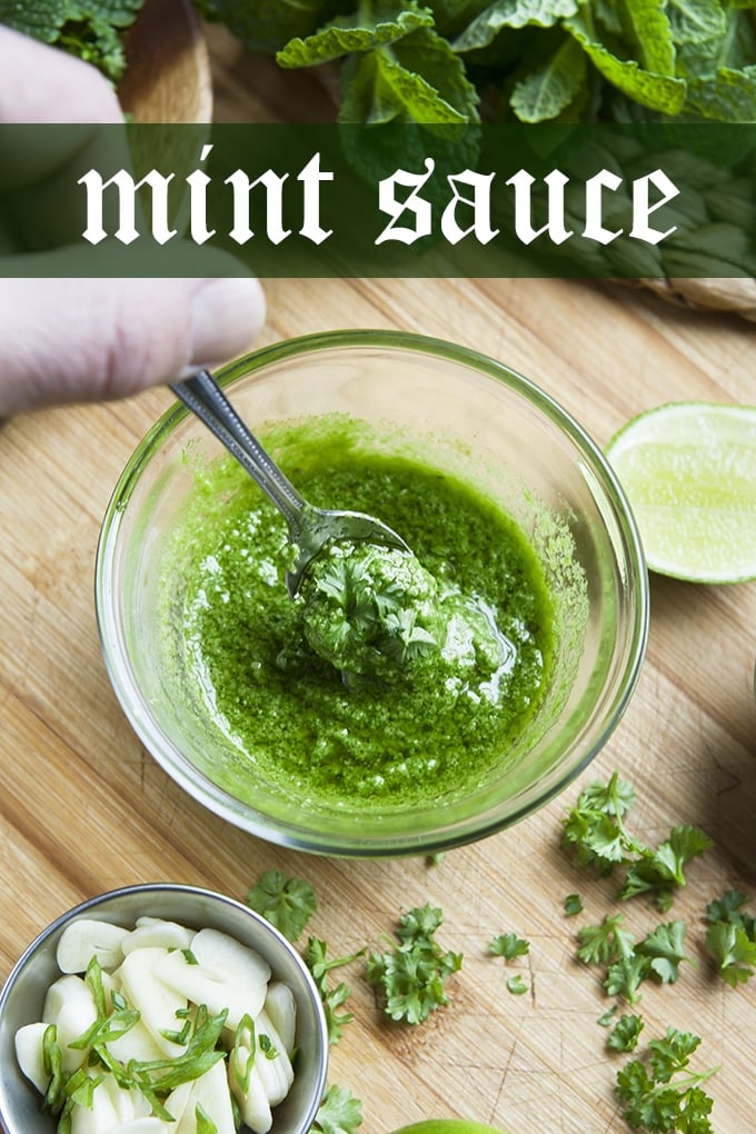 Mint sauce in a glass bowl with fresh mint leaves, lime, and sliced garlic nearby. There is also parlsey scattered on the wooden surface that the glass bowl is on. There's a spoon in the bowl and a hand holding the spoon.