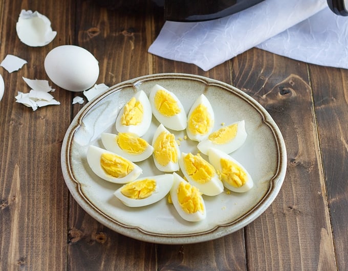 tan plate with brown line around edge with quartered hard boiled eggs on plate; white cloth in background as well as whole hard boiled egg with broken shell
