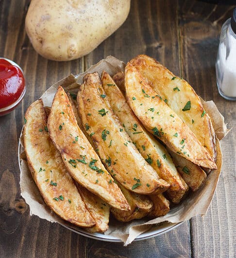 potato wedges garnished with parsley on paper lined dish with condiment bowl of ketchup behind it, salt shaker, and whole potato