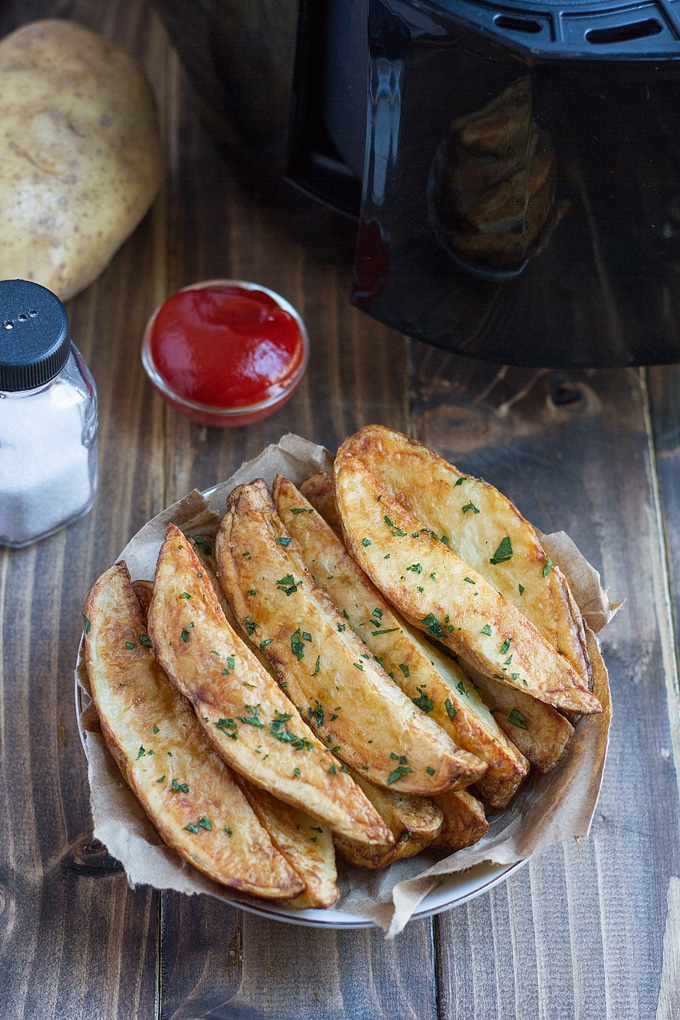 potato wedges garnished with parsley on paper lined dish with condiment bowl of ketchup behind it, salt shaker, and whole potato; air fryer in background