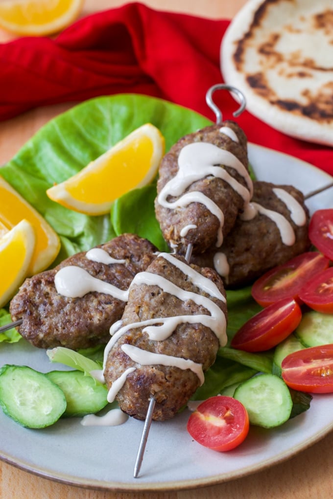 lamb kofta on a bed of lettuce served with lemon wedges, cucumber slices, tomato wedges, and drizzled with tahini sauce; pita and red cloth in background