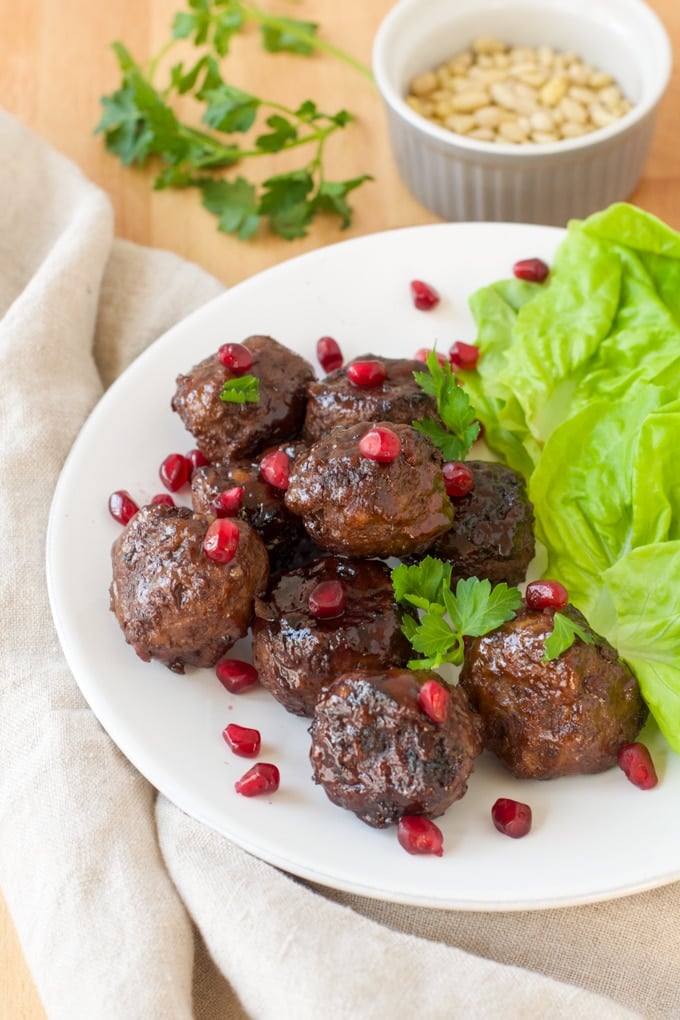 lamb meatballs garnished with pomegranate arils and green herbs; lettuce served on the side; condiment bowl of pine nuts in background