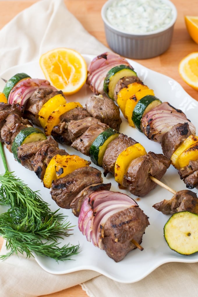 skewers with lamb, red onion, zucchini, and yellow bell pepper on them; lemon and tzatziki sauce in background