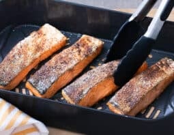 salmon filets in air fryer basket with tongs holding one piece; yellow and white striped cloth under basket