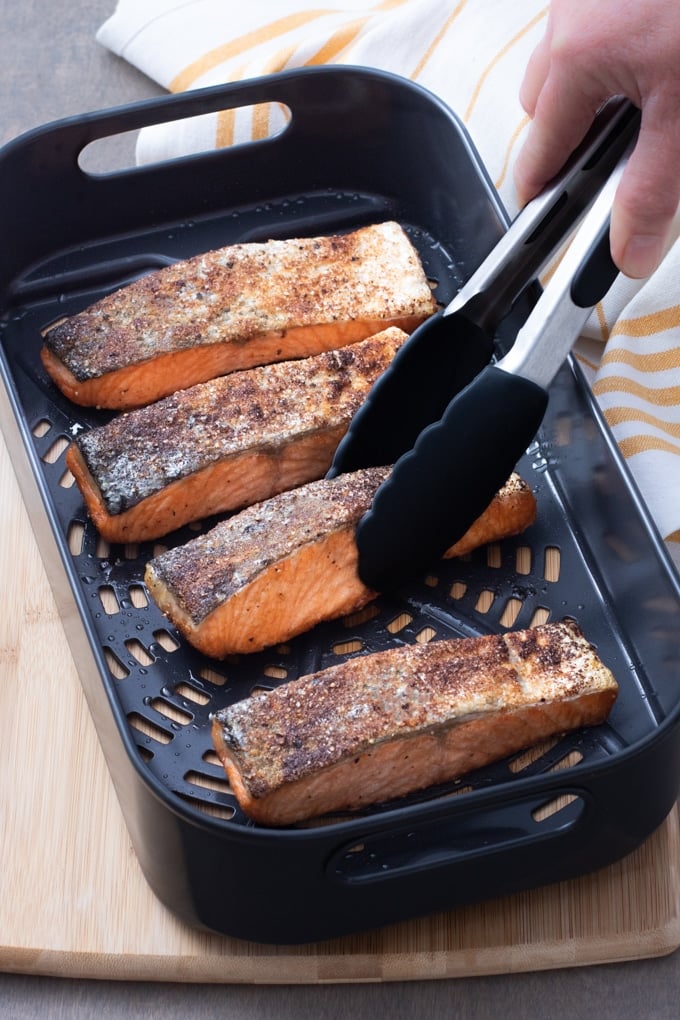 salmon filets in air fryer basket with tongs holding one piece; yellow and white striped cloth under basket