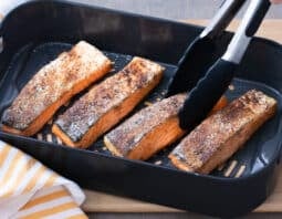 salmon filets in air fryer basket with tongs holding one piece; yellow and white striped cloth under basket