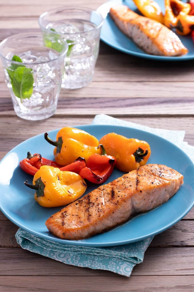 grilled seasoned salmon on light blue plate with grilled baby bell peppers; blue napkin under plate; glasses of water in background as well as second plate of same food