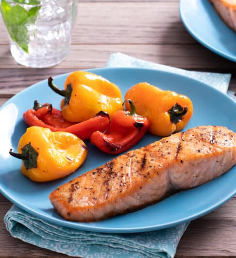grilled seasoned salmon on light blue plate with grilled baby bell peppers; blue napkin under plate; glasses of water in background