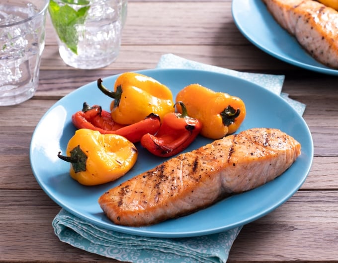 grilled seasoned salmon on light blue plate with grilled baby bell peppers; blue napkin under plate; glasses of water in background