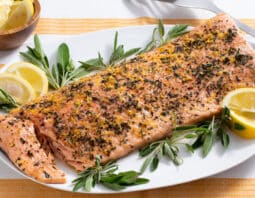 salmon crusted with sage, lemon zest and black pepper with sage and lemon wedge garnish on white platter with yellow an dwhite striped cloth under plate; small bowl of lemon wedges in background