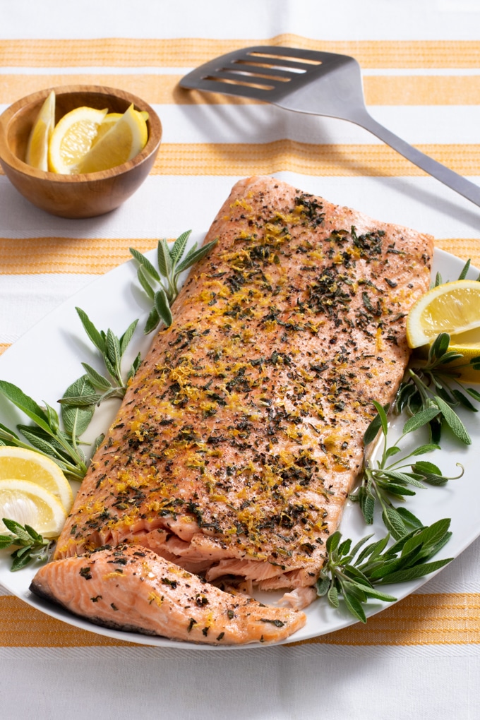 salmon crusted with sage, lemon zest and black pepper with sage and lemon wedge garnish on white platter with yellow an dwhite striped cloth under plate; small bowl of lemon wedges in background; spatula in background