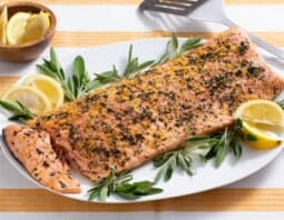 salmon crusted with sage, lemon zest and black pepper with sage and lemon wedge garnish on white platter with yellow an dwhite striped cloth under plate; small bowl of lemon wedges in background