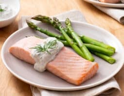 white plate topped with salmon fillet, topped with a lemon dill sauce and dill garnish; asparagus on plate; condiment dish with lemon dill sauce in bacjground