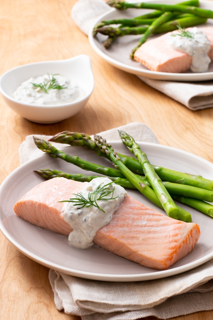 white plate topped with salmon fillet, topped with a lemon dill sauce and dill garnish; asparagus on plate; condiment dish with lemon dill sauce and another plate of the same food in background