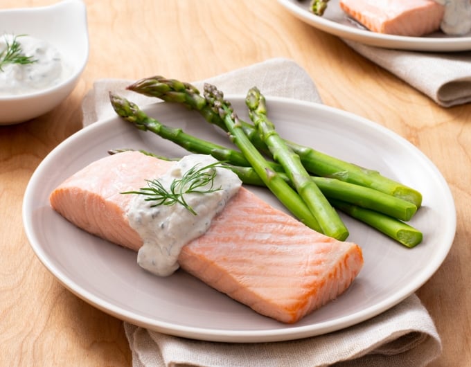 white plate topped with salmon fillet, topped with a lemon dill sauce and dill garnish; asparagus on plate; condiment dish with lemon dill sauce in background