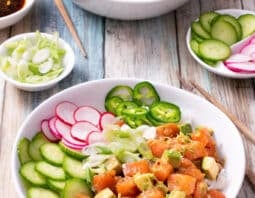 white bowl with cubed salmon, avocado, sliced cucumber, radished, and jalapenos on rice; chopsticks to right, small dish of sliced cucumber and radish in right hand corner; sliced green onion and soy sauce in condiment bowls to left; second bowl of food in background