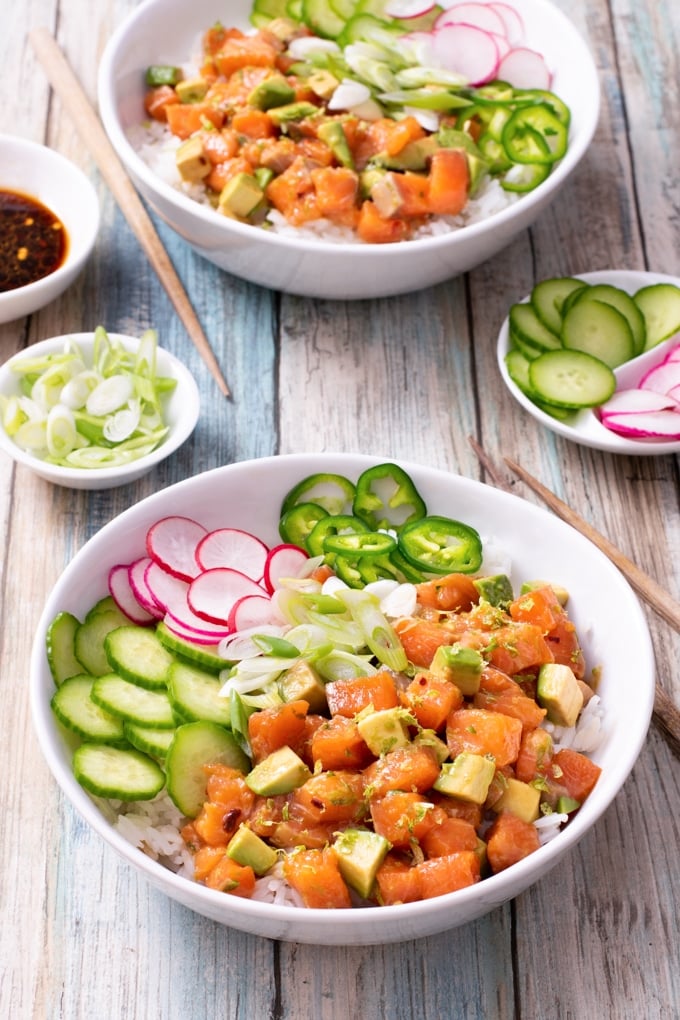 white bowl with cubed salmon, avocado, sliced cucumber, radished, and jalapenos on rice; chopsticks to right, small dish of sliced cucumber and radish in right hand corner; sliced green onion and soy sauce in condiment bowls to left; second bowl of food in background