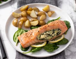 salmon topped with seasoning and stuffed with spinach and cream cheese laying on a bed of spinach and lemon slices; on a white plate with quartered and roasted small potatoes forks to left of plate, salt and pepper to right