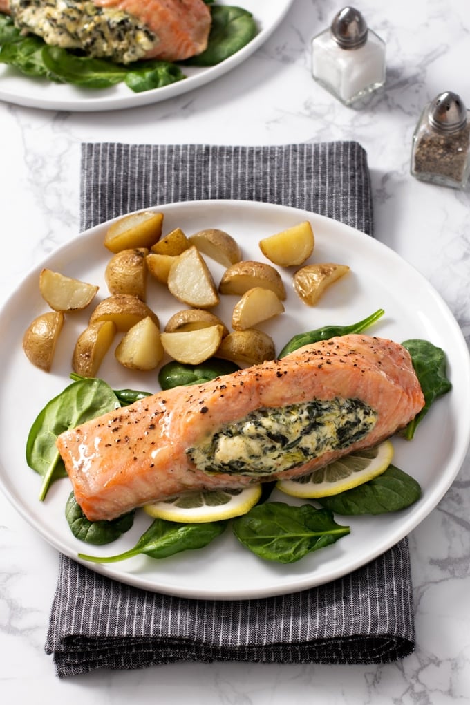 salmon topped with seasoning and stuffed with spinach and cream cheese laying on a bed of spinach and lemon slices; on a white plate with quartered and roasted small potatoes forks to left of plate, salt and pepper to right, second plate of food in top left corner