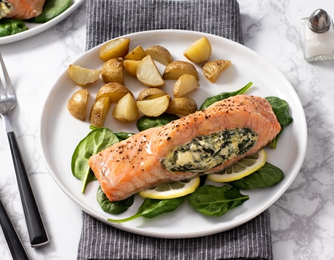 salmon topped with seasoning and stuffed with spinach and cream cheese laying on a bed of spinach and lemon slices; on a white plate with quartered and roasted small potatoes forks to left of plate, salt and pepper to right