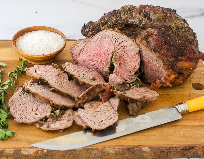boneless leg of lamb, half sliced with knife in front of it and fresh green herbs to left; small condiment bowl of salt behind it
