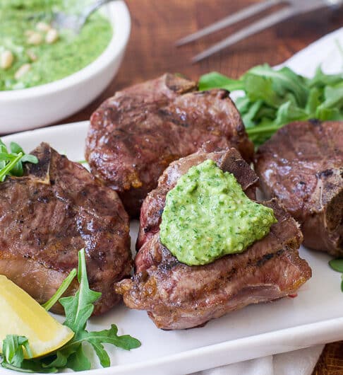 grilled lamb chops on bed of arugula on white platter garnished with green pesto and lemon wedges; condiment bowl of green pesto behind it