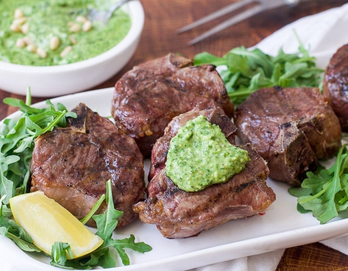 grilled lamb chops on bed of arugula on white platter garnished with green pesto and lemon wedges; condiment bowl of green pesto behind it