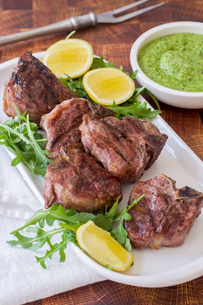 grilled lamb chops on bed of arugula on white platter garnished with green pesto and lemon wedges; condiment bowl of green pesto behind it