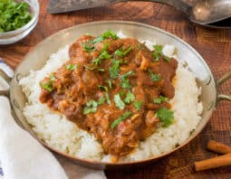 bowl with rice topped with indian lamb stew with an herb garnish; white and blue striped cloth under bowl; cinnamon stick and fresh green herb in background