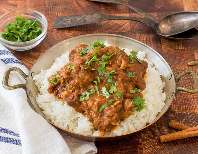 bowl with rice topped with indian lamb stew with an herb garnish; white and blue striped cloth under bowl; cinnamon stick and fresh green herb in background