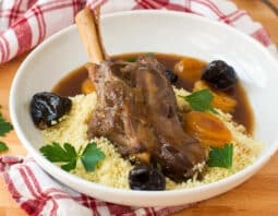 lamb shank served in white bowl over couscous with fresh herb garnish, prunes, apricots, and au jus