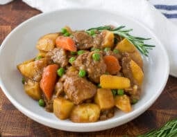 lamb, potato, carrots, peas in a white bowl with sprig of rosemary