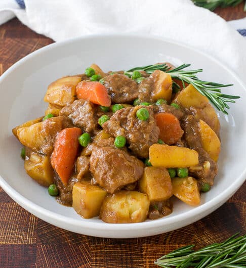 lamb, potatoe, carrots, and peas in a white bowl with a sprig of rosemary garnish