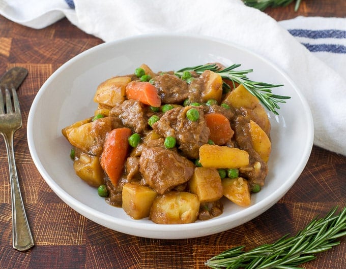lamb, potatoe, carrots, and peas in a white bowl with a sprig of rosemary garnish