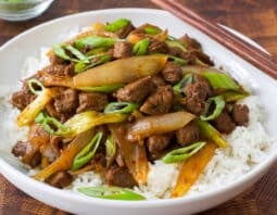 white bowl with rice topped with lamb and onion stir fry garnished with green scallion; chopsticks on edge of bowl; condiment bowl with sliced green scallion in background