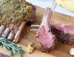 wooden board with garlic herb panko crusted rack of lamb and 2 chops; roasted garlic cloves and sprig of rosemary