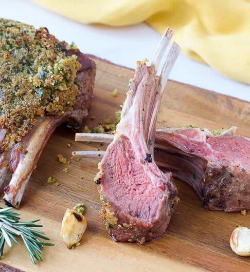 wooden board with garlic herb panko crusted rack of lamb and 2 chops; roasted garlic cloves and sprig of rosemary