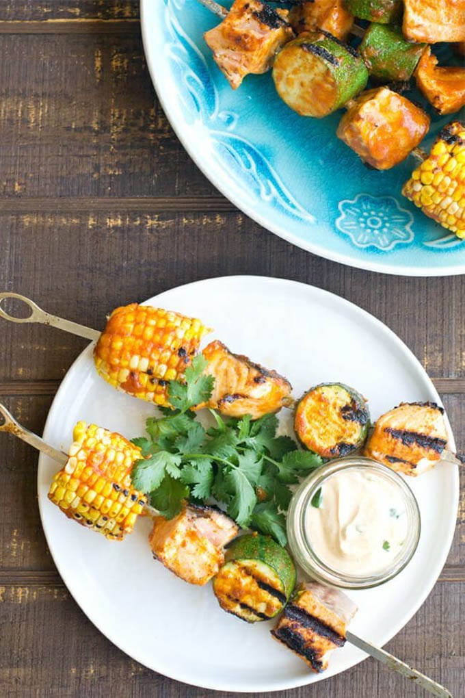 white plate with skewer with salmon, corn and zucchini on it; condiment bowl with sauce and herb garnish