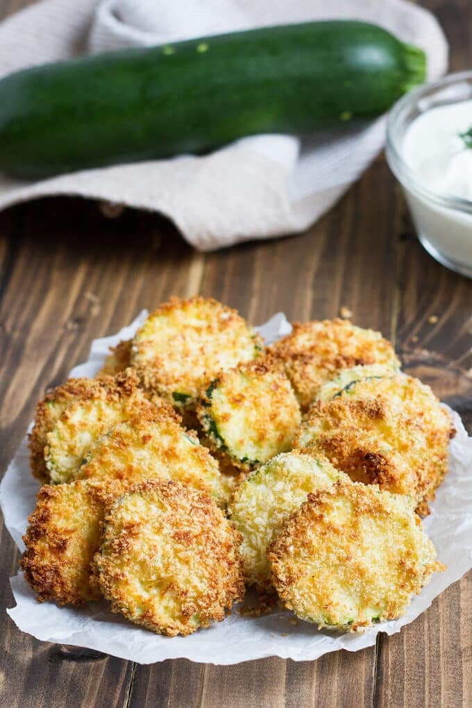 Round slices of zucchini coated in breadcrumbs that are golden brown from the air fryer. A fresh zucchini and a dipping sauce in a bowl are in the background.